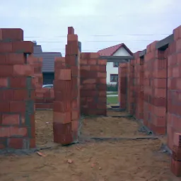 Wnętrze powstającego domu z czerwonej cegły, widoczne ściany działowe i otwory drzwiowe, w tle domy jednorodzinne